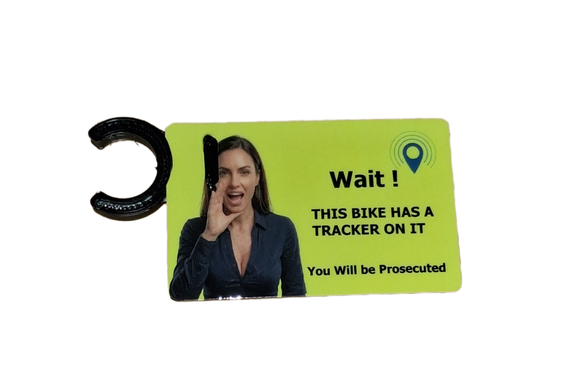 A warning tag with a bright lime green background, featuring a photo of a woman on a phone and the text 'Wait! THIS BIKE HAS A TRACKER ON IT You Will be Prosecuted'. The tag has a black plastic C-ring on the left side, set against a plain white background.