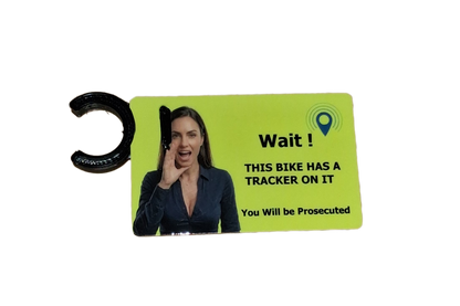A warning tag with a bright lime green background, featuring a photo of a woman on a phone and the text 'Wait! THIS BIKE HAS A TRACKER ON IT You Will be Prosecuted'. The tag has a black plastic C-ring on the left side, set against a plain white background.